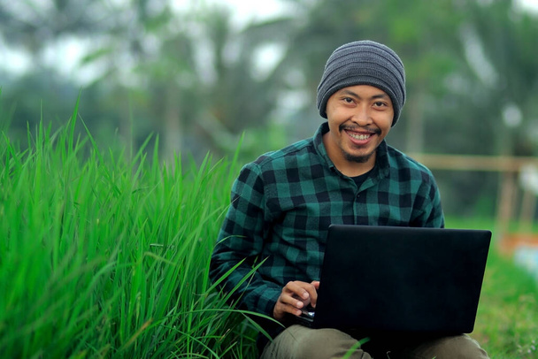 Indonesischer junger Mann mit Mütze und grünem Flanellhemd bei der Arbeit inmitten von Reisfeldern. - Foto, Bild