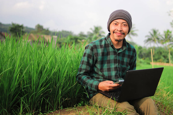 Jeune homme indonésien avec bonnet et chemise en flanelle verte travaillant à distance au milieu des champs de riz. - Photo, image