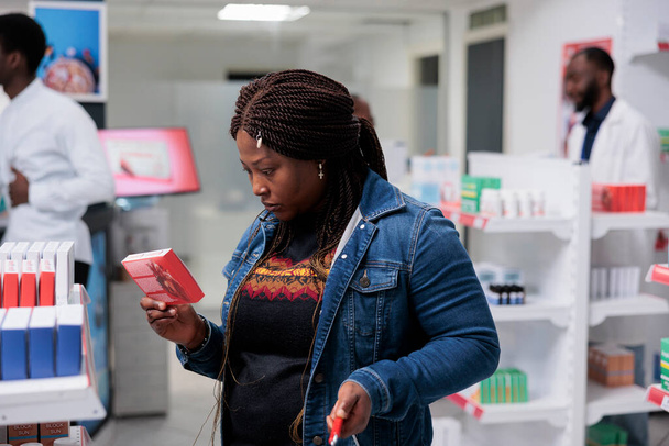 Africký zákazník kupující léky na srdce v lékárně, návod na čtení, balení tablet, stojící v lékárně. Klient vybírá vitamíny, střední střela - Fotografie, Obrázek