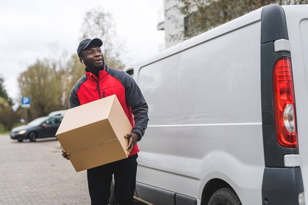 Young adult black delivery guy walking holding cardboard box parcel unloaded out of white van. Horizontal outdoor shot. High quality photo - Foto, Bild
