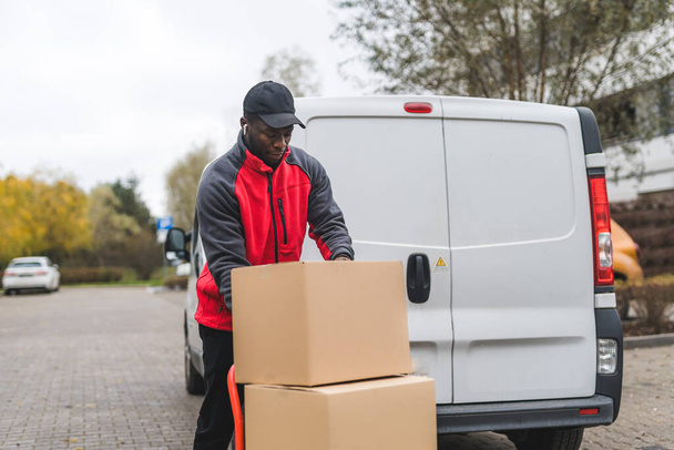 Package delivery concept. Black handsome male courier using hand truck to deliver big packages packed in cardboard boxes. White delivery van in the background. High quality photo - Фото, изображение