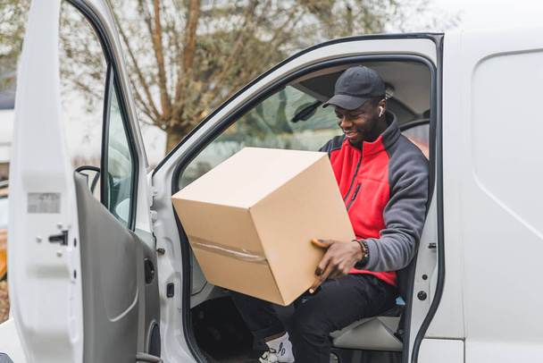 Black young adult delivery guy in work uniform sitting in door of white van looking at cardboard box parcel. Horizontal outdoor shot. High quality photo - 写真・画像