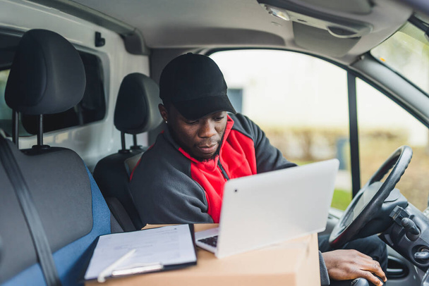 In-car view of busy focused Black middle-aged delivery man in uniform using his silver laptop to cross out next parcels. The use of vehicle - white van. High quality photo - Foto, afbeelding