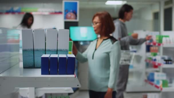 Donna asiatica alla ricerca di farmaci in farmacia, guardando scaffali pieni di scatole di pillole e bottiglie. Pacchi medici di lettura del cliente della farmacia, prodotti farmaceutici. - Filmati, video