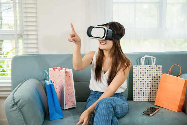 Happy young woman customer wearing 3d VR glasses virtual reality headset with shopping bags around she touching and pointing on air to online shopping in living room at home, digital cyberspace - Foto, afbeelding