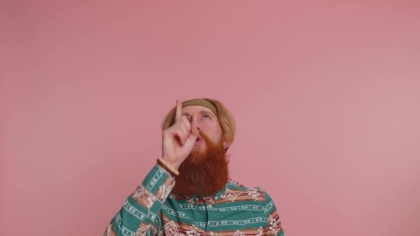 Hippie rudy brodaty mężczyzna we wzorzystej koszuli pokazuje kciuki do góry i wskazując nad głową na pustej przestrzeni. Miejsce na logo reklamowe, baner, tekst. Hipster rudy facet na różowym tle - Materiał filmowy, wideo