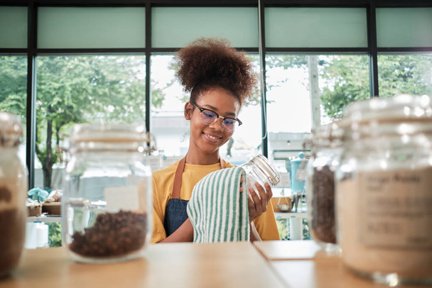Egy fiatal fekete női boltos üvegedényeket tisztít természetes biotermékekből újrahasznosítható tartályokban egy utántöltő boltban, nulla hulladék, egy műanyagmentes élelmiszerbolt és egy környezetbarát kiskereskedelmi vállalkozás.. - Fotó, kép