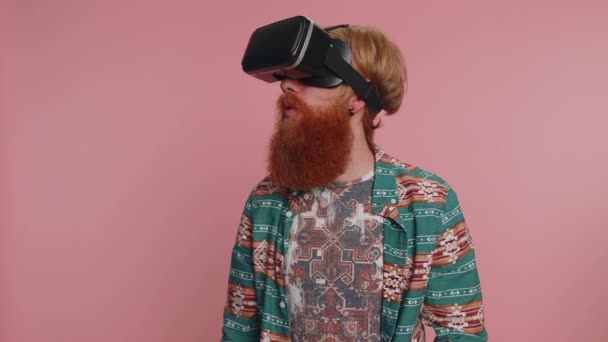 Redhead bearded hippie man using headset helmet app to play simulation innovation game. Watching virtual reality 3D 360 video. Young ginger guy boy in VR goggles isolated on pink studio background - Footage, Video