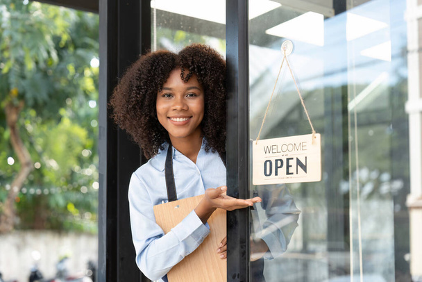 Malá obchodní africká majitelka se usmívá a otáčí cedulí pro otevření kavárny. Happy afro-americká servírka podnikatel v zástěře současné znamení na otevření dveří. - Fotografie, Obrázek