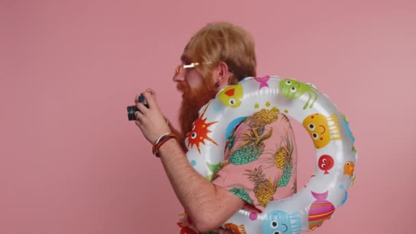 レトロなカメラで写真を撮るサングラスの幸せなヒップスター髭の男の観光客の写真家。旅行、夏休み。Hippie redhead smiling guy traveler on studio pink background - 映像、動画