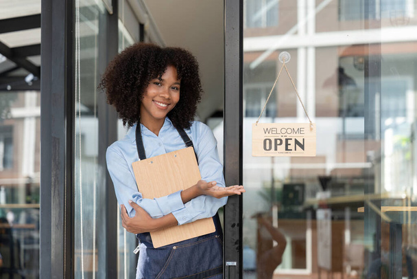 Small business african female owner smiling while turning sign for opening of cafe. Happy afro-american waitress entrepreneur in apron present sign on door opening. - Photo, Image