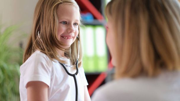 Portrait of cheerful small girl using medical stethoscope and looking at doctor with happiness. Healthcare, medicine and childhood concept - Photo, Image