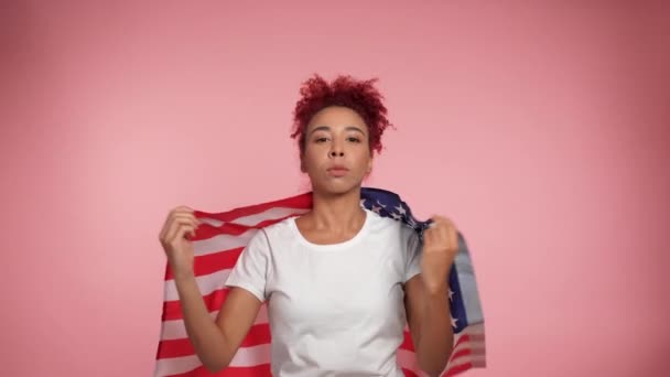 Komoly afro-amerikai hazafi vörös hajú göndör nő függönyök amerikai zászló felett vállak néz kamera. Portré női álló USA zászló izolált rózsaszín háttér. Július 4. Amerika függetlenségének napja - Felvétel, videó
