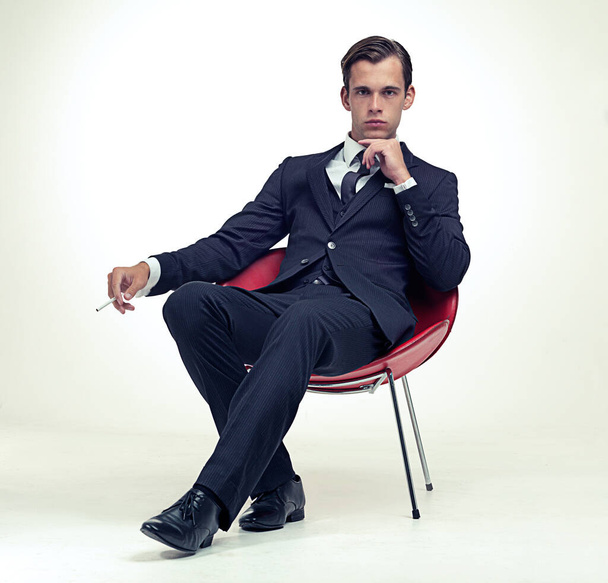 Savuava menestys. Studio muotokuva komea nuori mies liituraita puku istuu tuolilla ja tupakointi - Valokuva, kuva