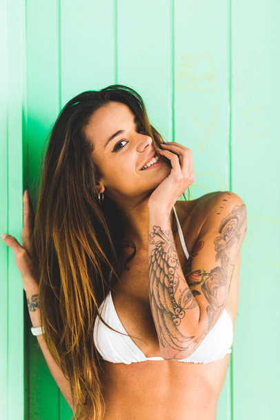 Portrait of a young adult with tattooed body standing against a green door at a beach wearing a bikini - Foto, Imagen