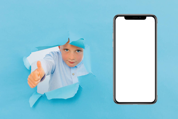 Ευτυχισμένο χαριτωμένο αγόρι έχει τη διασκέδαση παίζεται σε μπλε φόντο τοίχο, σκαρφαλώνει μέσα από μια τρύπα στην εφημερίδα κοντά στο τεράστιο κινητό τηλέφωνο. Φωτεινά και αστεία συναισθήματα του αγοριού - Φωτογραφία, εικόνα