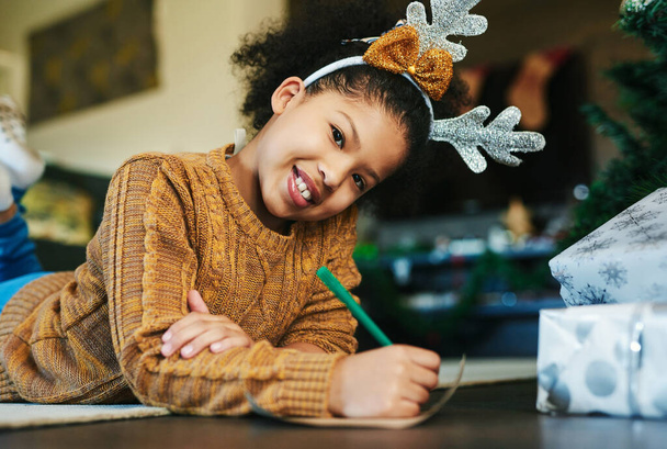 Boże Narodzenie, szczęście i dziewczyna pisząc listę do Świętego Mikołaja na prezent, prezenty i zabawki dla tradycji wakacyjnej. Festiwal, uroczystość i portret dziecka na podłodze do napisania listu w domu rodzinnym. - Zdjęcie, obraz