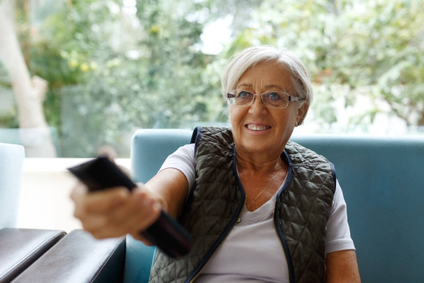 Joyeux sourire dame âgée avec les cheveux gris assis sur le canapé bleu dans le salon, pointant une télécommande de télévision pour changer de chaînes, excité de regarder des séries en ligne - Photo, image