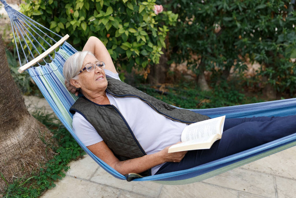 Sonriente mujer de pelo gris de buen aspecto con gafas mientras lee en hamaca en el jardín de verano, sonriendo - Foto, Imagen