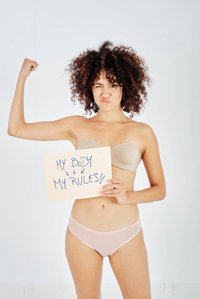 Латиноамериканська жіноча модель в нижній білизні з моїм тілом мої правила плакат підморгує і показує м'язи руки на сірому тлі - Фото, зображення