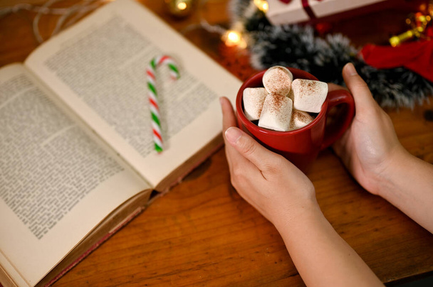 Une main féminine tenant une tasse de chocolat chaud avec guimauve sur une table en bois avec livre vintage et décor de Noël. - Photo, image