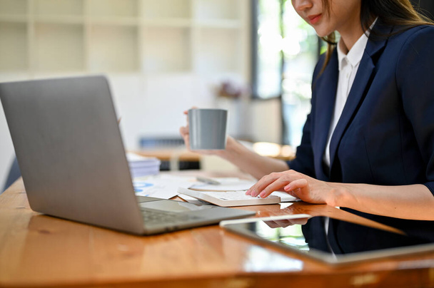 プロのアジアの女性会計士やビジネスマンは、彼女の机で作業しながらコーヒーを飲みながら、計算機を使用して販売報告書に取り組んでいます。クロップドとサイドビュー - 写真・画像