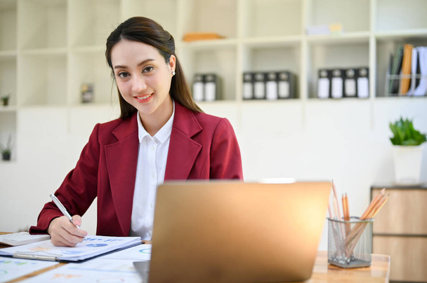 Attractive and charming young Asian businesswoman in stylish business suit sitting at her desk, working on document or report, smiling and looking at camera. - Foto, Bild