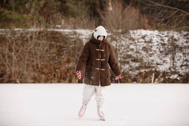 Gyermek korcsolyázás a befagyott tavon a havas erdőben. Egy kislány meleg ruhában és rózsaszín korcsolyában a jégpályán a hideg téli napon. Kellemes nyaralást!. - Fotó, kép
