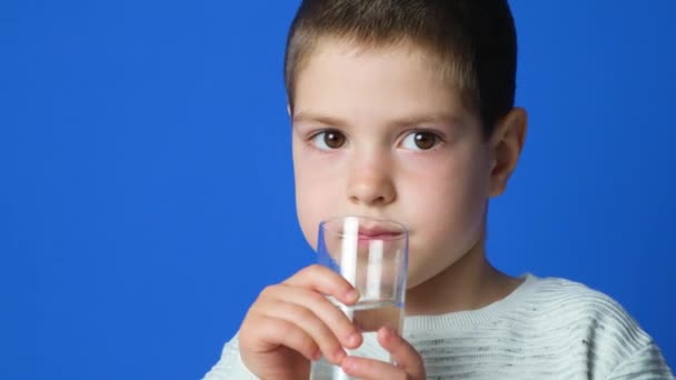 Niño preescolar feliz bebiendo agua de un vaso sobre un fondo azul. - Metraje, vídeo