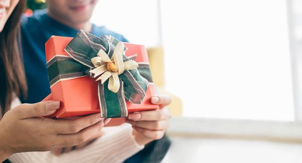 A closeup Young Asian woman hands hold a Christmas gift in a red box with ribbon with her boyfriend in a living room with a decorated Christmas tree in the background. - 写真・画像