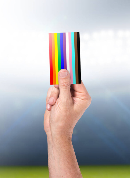 Ein männlicher Sportschiedsrichter oder Schiedsrichter hält eine LGBTQ-Farbkarte vor dem Hintergrund eines Sportstadions in die Luft - Foto, Bild