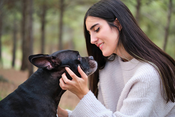 Young caucasian woman stroking her american staffordshire and french bulldog mixed breed dog at a pine forest. - Photo, Image