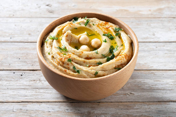 Chickpea hummus in a wooden bowl garnished with parsley, paprika and olive oil on wooden table - Fotografie, Obrázek