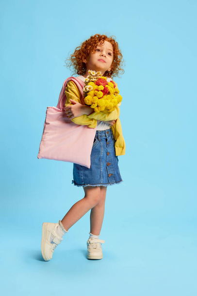 Portrait of cute stylish little girl, child with curly red hair posing with flowers isolated over blue background. Concept of childhood, emotions, lifestyle, fashion, happiness. Copy space for ad - Zdjęcie, obraz