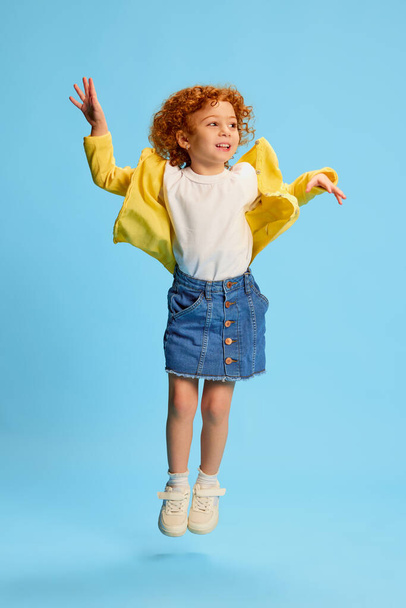 Portrait of cute little girl, child with curly red hair posing in stylish clothes isolated over blue background. Concept of childhood, emotions, lifestyle, fashion, happiness. Copy space for ad - Foto, afbeelding