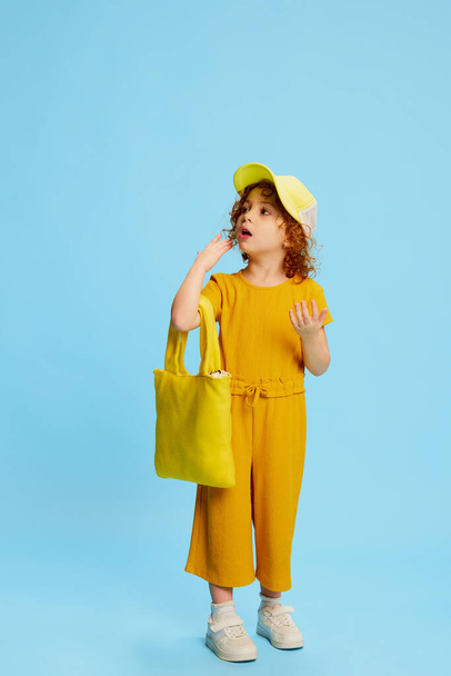Portrait of cute little girl, child with curly red hair posing, going shopping isolated over blue background. Concept of childhood, emotions, lifestyle, fashion, happiness. Copy space for ad - Фото, изображение
