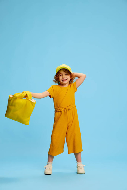 Portrait of cute little girl, child with curly red hair posing isolated over blue background. Stylish yellow outfit. Concept of childhood, emotions, lifestyle, fashion, happiness. Copy space for ad - Фото, изображение