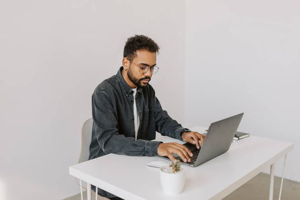 Foto lateral de hombre africano feliz sonriente en camisa gris con estilo con pelo rizado y gafas que trabajan en el ordenador portátil y mecanografía mientras está sentado en el escritorio blanco en el estudio de oficina moderno espacioso acogedor 4k. - Foto, imagen