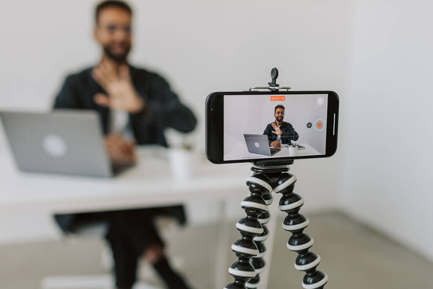 Μέτριο πλάνο του μαύρου άνδρα μέσω εστιασμένη κάμερα τηλέφωνο σε τρίποδο και κάθεται στο γραφείο με φορητό υπολογιστή και εξηγώντας κάτι στην κάμερα, ενώ μαγνητοσκόπηση vlog στο άνετο γραφείο στούντιο. Υψηλής ποιότητας υλικό - Φωτογραφία, εικόνα