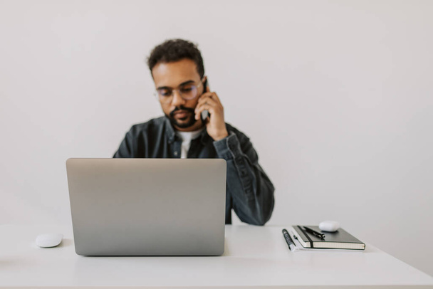 Serious black diverse male in grey shirt with eyeglasses curly hair talking on cell phone with colleagues sitting at the white clean desk in cozy stylish home office in front of laptop - Photo, image