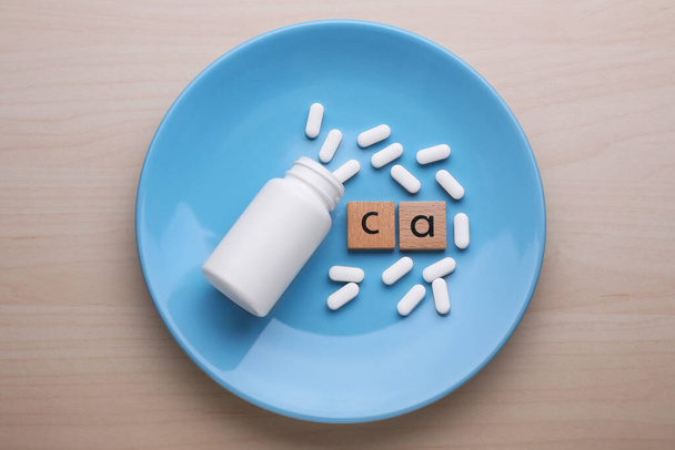 Wooden cubes with symbol Ca (Calcium), medical bottle and pills on light blue plate, top view - Photo, image