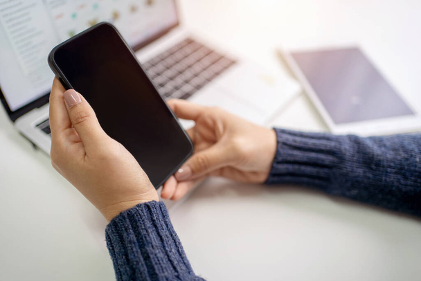 Mujer está utilizando un teléfono inteligente con una pantalla en blanco y un ordenador portátil en una mesa cercana. Ella está sentada en la oficina usando un suéter. - Foto, imagen