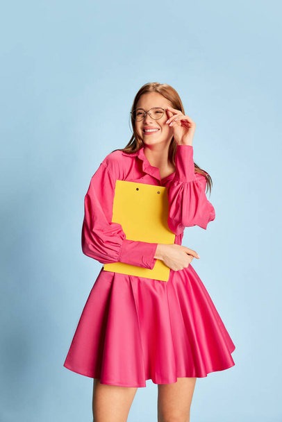 Porträt eines jungen schönen Mädchens in stilvollem rosa Kleid und Brille lächelnd, posierend isoliert vor blauem Hintergrund. Studentenleben. Konzept der Jugend, Schönheit, Mode, Emotionen, Mimik. Anzeige - Foto, Bild