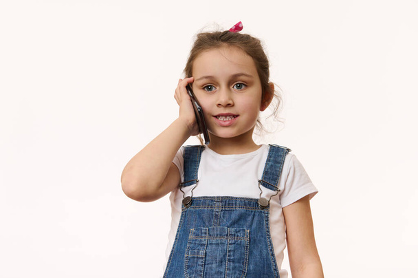 Entzückendes kaukasisches kleines Mädchen, in blauen Jeans-Overalls und weißem T-Shirt, telefonierend, isoliert vor weißem Hintergrund. Kopieren Sie Werbeflächen. Kinder und elektronische Digitaltechnik - Foto, Bild