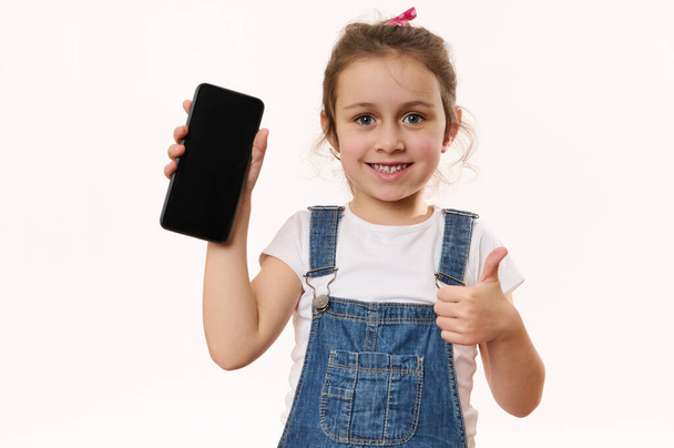 Bella bambina, carina bambina con smartphone in mano con schermo bianco con pubblicità spazio libero per il testo promozionale, sorridente con sorriso dentato, mostrando il pollice alla fotocamera. Bambini e gadget digitali - Foto, immagini