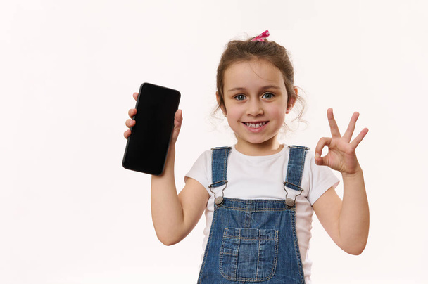 Lovely little kid, cute girl holding cell phone with blank screen with copy of advertising space for promo text, smiling with toothy smile, showing OK sign to camera. Children and digital gadgets - Фото, изображение