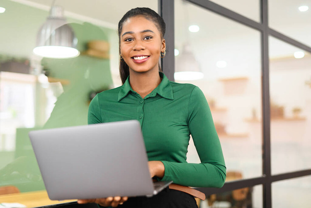 African-American businesswoman in smart casual wear standing in modern coworking space and holding laptop, female freelancer or student with laptop computer indoors smiling cheerfully - Foto, afbeelding