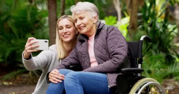 Vanhempi nainen, tytär ja selfie ulkona puistossa vanhusten pyörätuolissa kuntoutusta ja ilmaa saattohoito tai hoitaja. Aikuinen. lapsi ja vanha äiti yhdessä puhelimen kanssa kuvan saamiseksi Yhdistyneessä kuningaskunnassa. - Materiaali, video