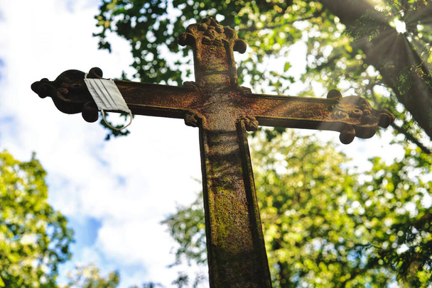Una croce di metallo arrugginito con una maschera appesa su di esso in un vecchio cimitero. Bei raggi di sole lo illuminano. - Foto, immagini