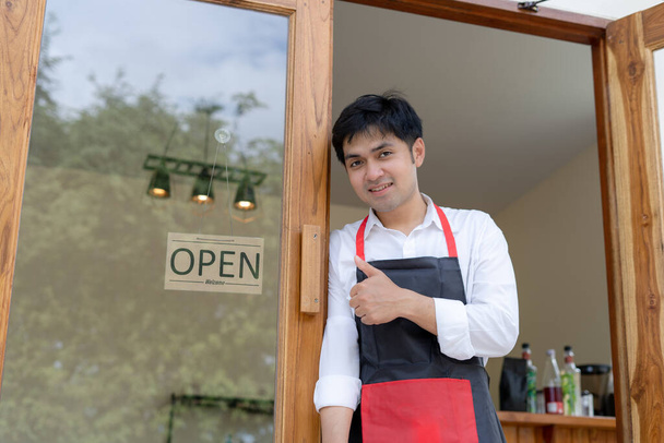Business owner thumbs up to show his readiness to welcome customers. waitress man turning on open sign board on glass door in modern cafe coffee shop. New normal, small business, cafe, life style. - Photo, Image
